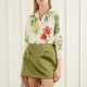 A woman wearing a floral top (white, red, green, and yellow) and olive green skirt