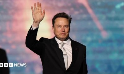 SpaceX chief executive Elon Musk walking onto a stage and waving, wearing a black suit, white collared shirt and shiny off-white neck tie.
