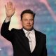 SpaceX chief executive Elon Musk walking onto a stage and waving, wearing a black suit, white collared shirt and shiny off-white neck tie.
