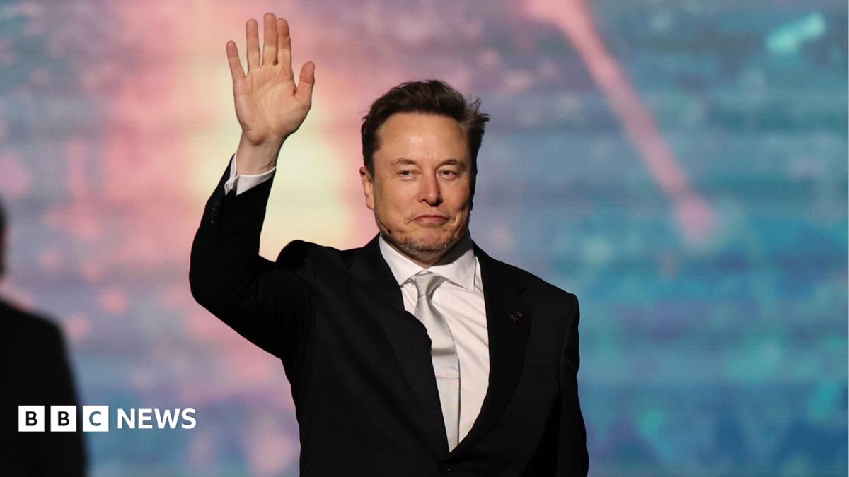 SpaceX chief executive Elon Musk walking onto a stage and waving, wearing a black suit, white collared shirt and shiny off-white neck tie.