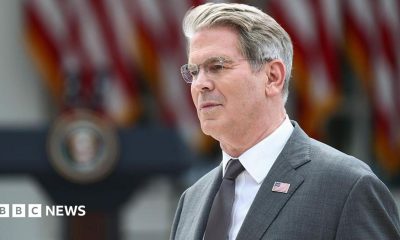 US Treasury Secretary Scott Bessent's head and shoulders in profile. He has grey hair and glasses. He is wearing a grey suit with a US flag lapel pin, a silver tie, and a white shirt. Behind him in soft focus are US flags and a podium.