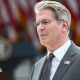 US Treasury Secretary Scott Bessent's head and shoulders in profile. He has grey hair and glasses. He is wearing a grey suit with a US flag lapel pin, a silver tie, and a white shirt. Behind him in soft focus are US flags and a podium.