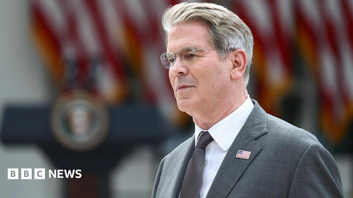 US Treasury Secretary Scott Bessent's head and shoulders in profile. He has grey hair and glasses. He is wearing a grey suit with a US flag lapel pin, a silver tie, and a white shirt. Behind him in soft focus are US flags and a podium.