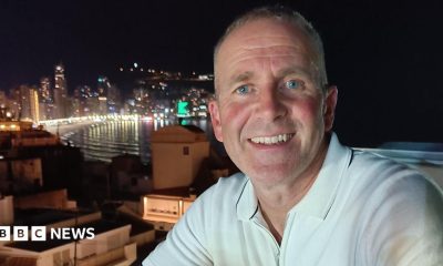Les Howard, who is wearing a white polo shirt, is starring directly at the camera and is smiling. There is a city skyline in the background.