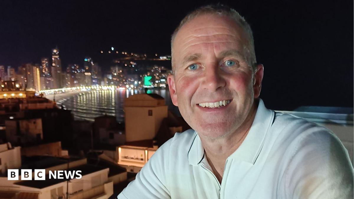 Les Howard, who is wearing a white polo shirt, is starring directly at the camera and is smiling. There is a city skyline in the background.