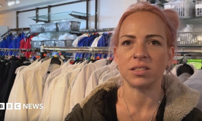 A woman, aged in her 20s or 30s, with pink dyed hair and wearing a green coat with a fur lined hood, speaks into the camera. She has a nose ring and a necklace, and is standing in front of a clothes rail lined with white school shirts.