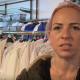 A woman, aged in her 20s or 30s, with pink dyed hair and wearing a green coat with a fur lined hood, speaks into the camera. She has a nose ring and a necklace, and is standing in front of a clothes rail lined with white school shirts.