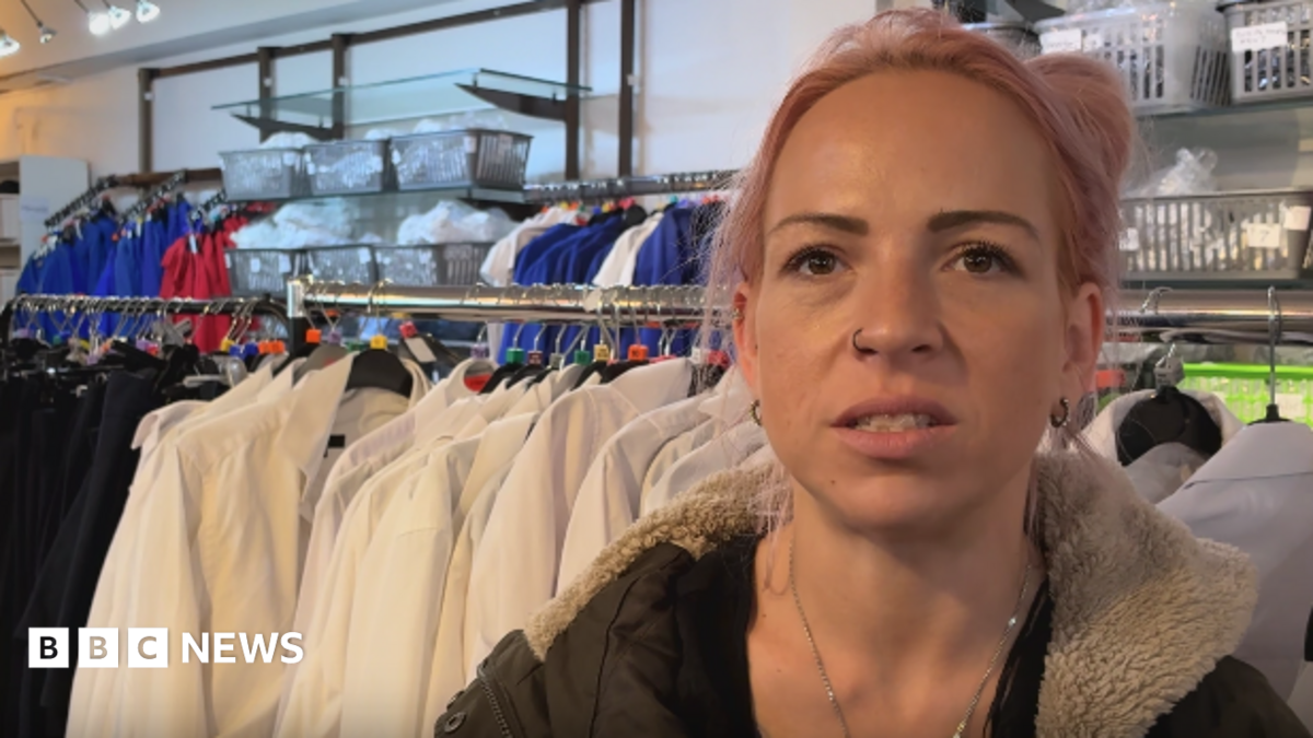 A woman, aged in her 20s or 30s, with pink dyed hair and wearing a green coat with a fur lined hood, speaks into the camera. She has a nose ring and a necklace, and is standing in front of a clothes rail lined with white school shirts.