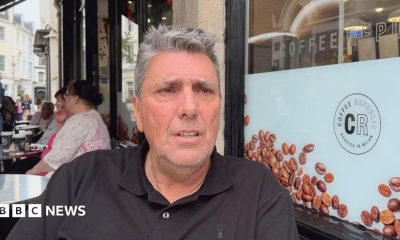 A person wearing a black polo shirt sits at an outdoor café table beside a large window displaying coffee bean graphics and the text “Coffee Republic” and “Roasted in Milan.” Several takeaway coffee cups are visible on nearby tables, with other café patrons seated in the background along a pedestrian street lined with buildings. The scene is photographed in daylight, with the seated person in sharp focus and the café surroundings clearly visible.