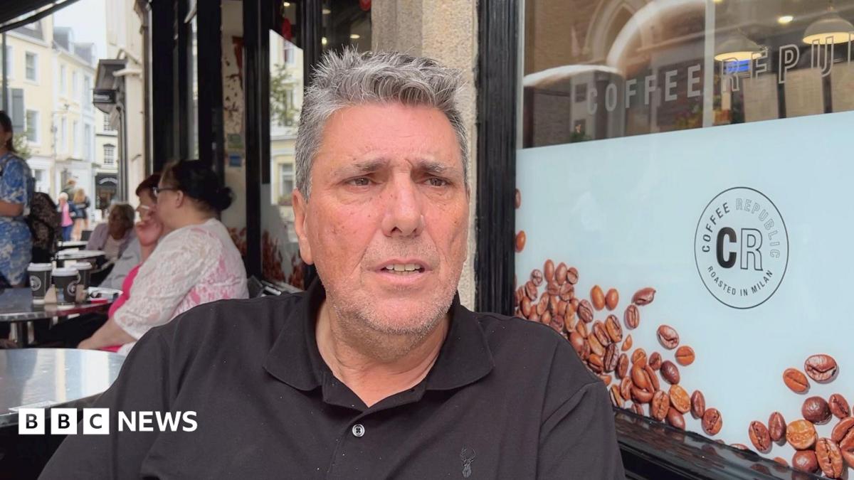 A person wearing a black polo shirt sits at an outdoor café table beside a large window displaying coffee bean graphics and the text “Coffee Republic” and “Roasted in Milan.” Several takeaway coffee cups are visible on nearby tables, with other café patrons seated in the background along a pedestrian street lined with buildings. The scene is photographed in daylight, with the seated person in sharp focus and the café surroundings clearly visible.