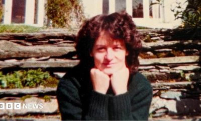 A woman sits looking at the camera with her chin resting on her balled fists. She is slightly smiling. She has curly dark brown hair and is wearing a dark green jumper. Behind her is a stone wall with some green plants growing out of it and behind that is a white house.