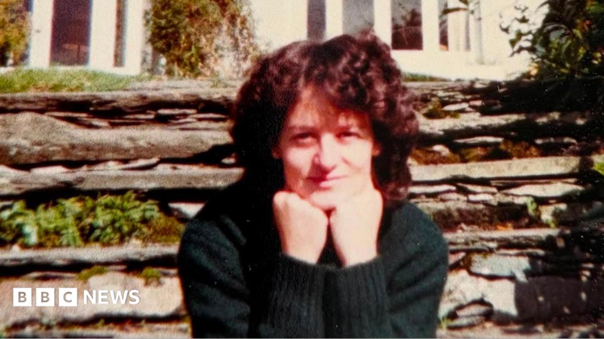 A woman sits looking at the camera with her chin resting on her balled fists. She is slightly smiling. She has curly dark brown hair and is wearing a dark green jumper. Behind her is a stone wall with some green plants growing out of it and behind that is a white house.