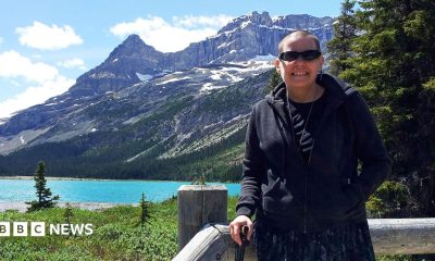 Cindy Baldassi is in the foreground wearing sunglasses and a dark hoody and navy blue dress. She is looking at the camera and behind her is a beautiful blue lake and towering mountain peaks with snow on top of them.