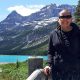 Cindy Baldassi is in the foreground wearing sunglasses and a dark hoody and navy blue dress. She is looking at the camera and behind her is a beautiful blue lake and towering mountain peaks with snow on top of them.