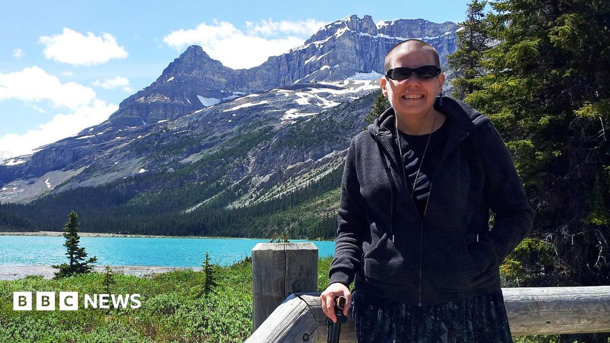 Cindy Baldassi is in the foreground wearing sunglasses and a dark hoody and navy blue dress. She is looking at the camera and behind her is a beautiful blue lake and towering mountain peaks with snow on top of them.