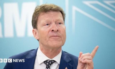 Richard Tice, in blue jacket, white shirt, and black and white tie, points with his finger while talking to reporters