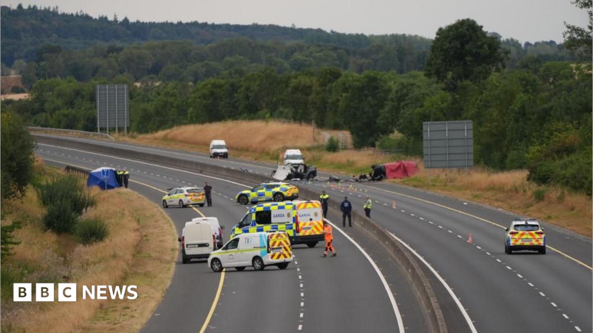 A wide shot of a car crash site. Many emergency service vehicles are on the road and debris from the crash.