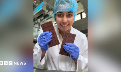 A woman holding two bars of chocolate while smiling into the camera. She is wearing a white gown with purple gloves and a blue hair net.