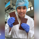 A woman holding two bars of chocolate while smiling into the camera. She is wearing a white gown with purple gloves and a blue hair net.