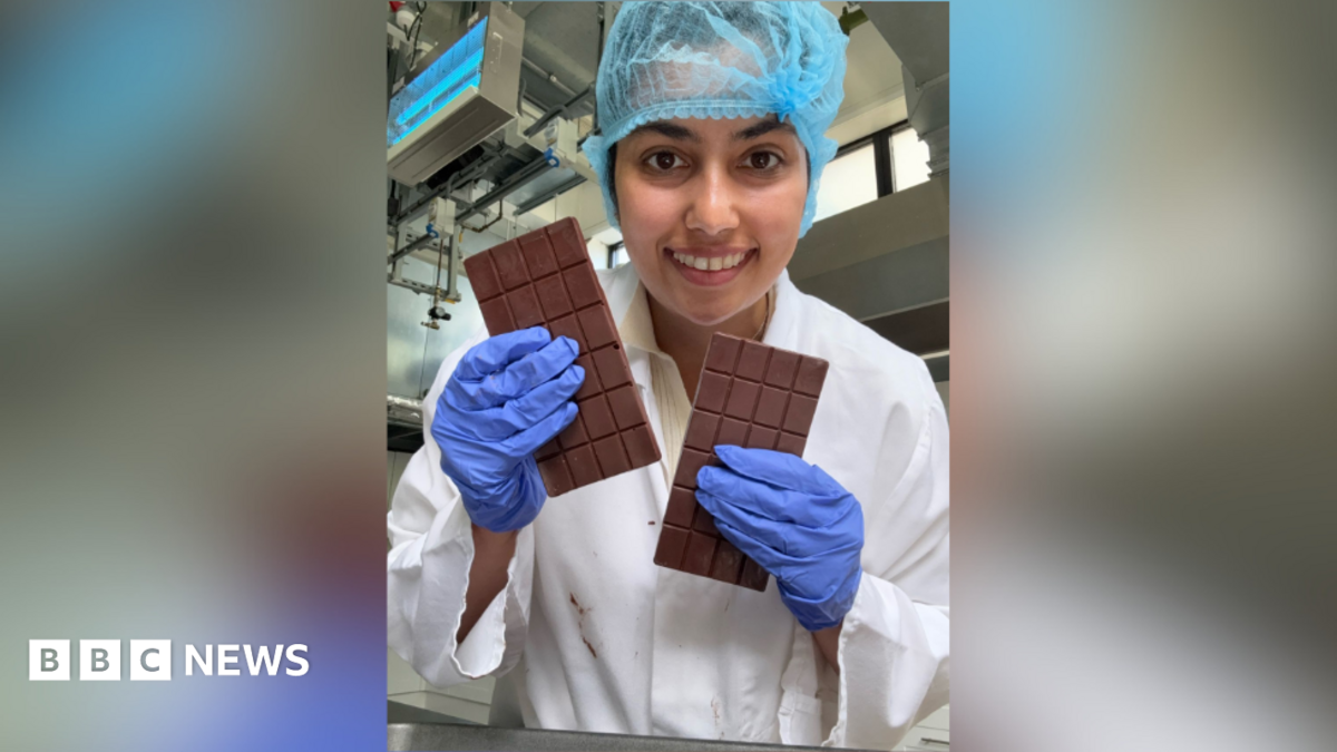 A woman holding two bars of chocolate while smiling into the camera. She is wearing a white gown with purple gloves and a blue hair net.
