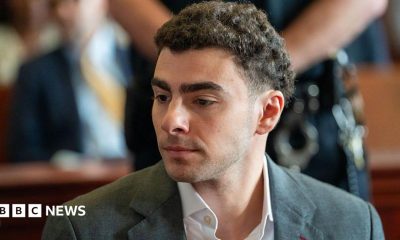 Luigi Mangione is seen from the shoulders up, sitting at the defence table in court. He wears a charcoal grey suit.