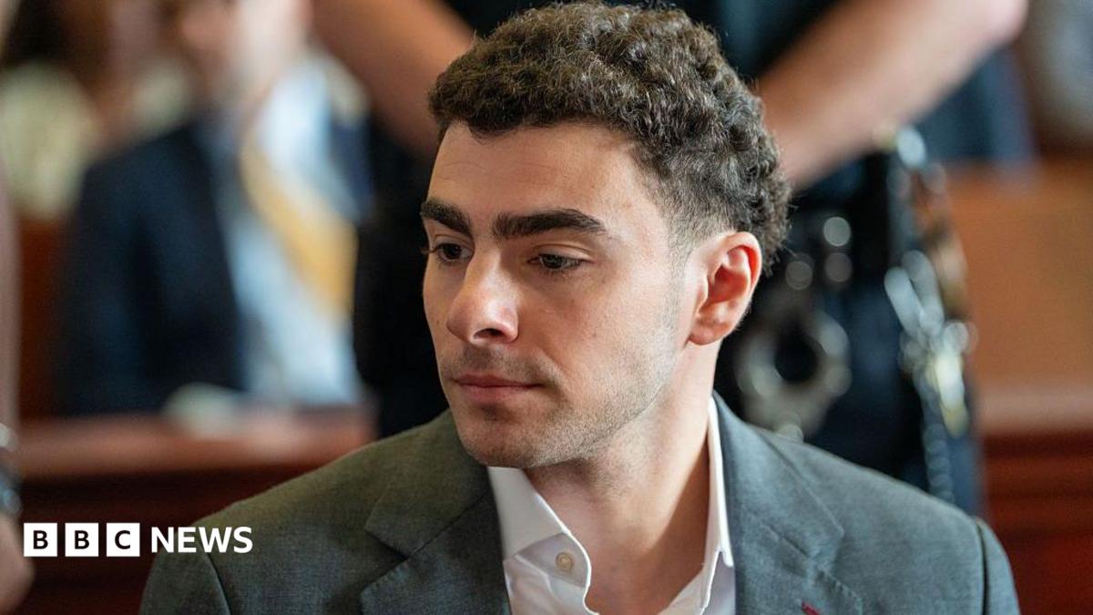 Luigi Mangione is seen from the shoulders up, sitting at the defence table in court. He wears a charcoal grey suit.