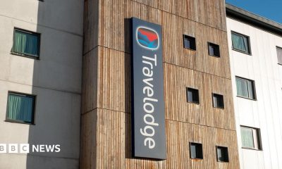A man in a blue T-shirt, arms crossed, looking serious, standing outside a Travelodge hotel.