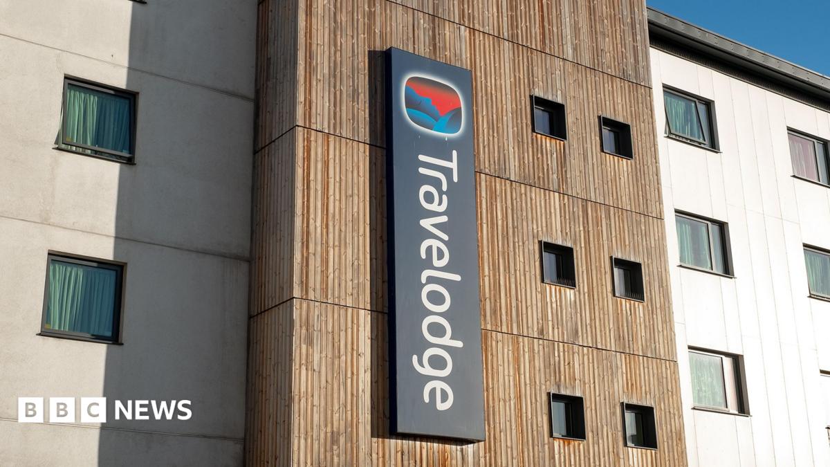 A man in a blue T-shirt, arms crossed, looking serious, standing outside a Travelodge hotel.