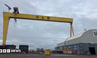 A wide shot of a yellow ship building crane with the letters H&W in back bold writing. A large grey shed with a curved roof is on the right hand side of the image.