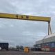 A wide shot of a yellow ship building crane with the letters H&W in back bold writing. A large grey shed with a curved roof is on the right hand side of the image.