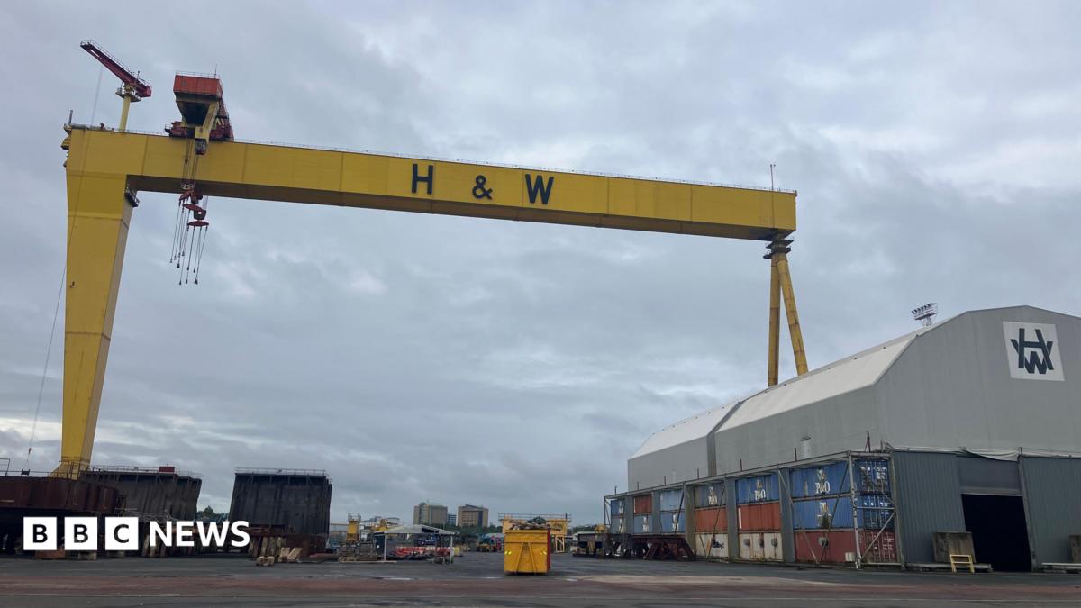 A wide shot of a yellow ship building crane with the letters H&W in back bold writing. A large grey shed with a curved roof is on the right hand side of the image.