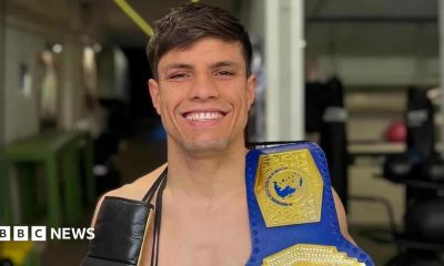 A man with dark hair smiling with a boxing belt over his shoulder.