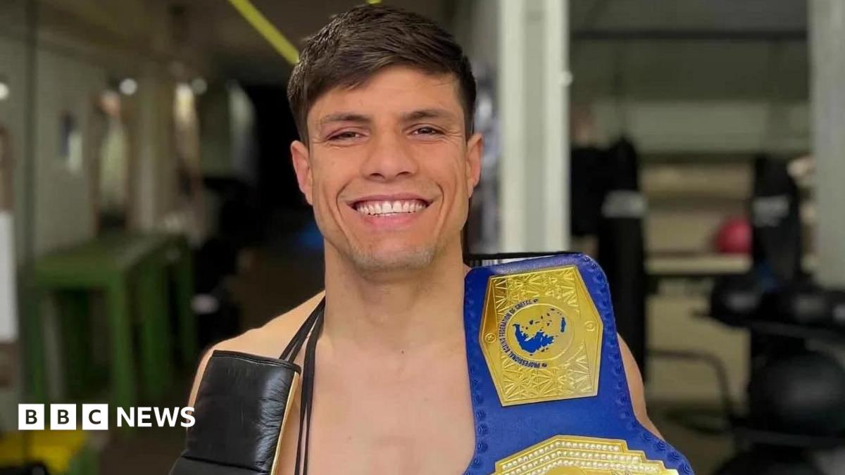 A man with dark hair smiling with a boxing belt over his shoulder.