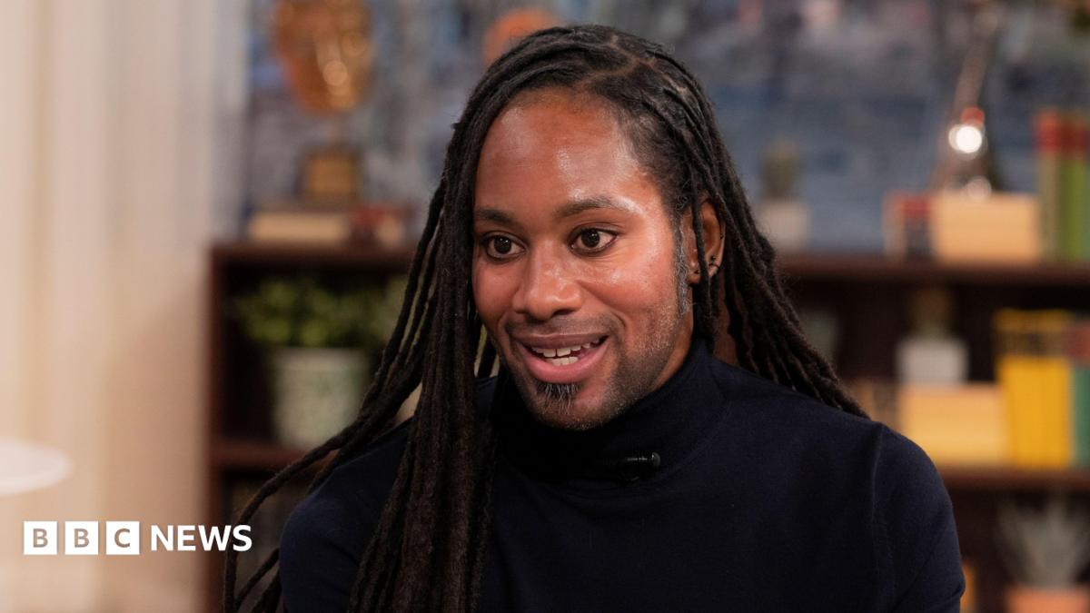 Jason Arday pictured during a TV interview in March 2023. He wears a black turtle-neck top.