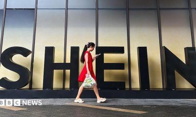 A woman in a red dress looks at her smartphone while walking past a Shein sign outside the company's office in Guangzhou, China.
