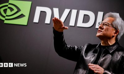 Nvidia CEO Jensen Huang standing in front of a screen with the company's name and logo, gesturing upward with his hands, wearing a black leather jacket over a black shirt.