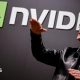 Nvidia CEO Jensen Huang standing in front of a screen with the company's name and logo, gesturing upward with his hands, wearing a black leather jacket over a black shirt.