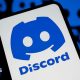 The blue Discord logo on a phone against a white background. In the background are more Discord logos on a black background.