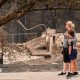 A woman dressed in black capris, grey jumper and black trousers wraps her arm around a young boy in black shorts, black top and black trainers, as both survey the damage of a burnt tree and rubble of a building