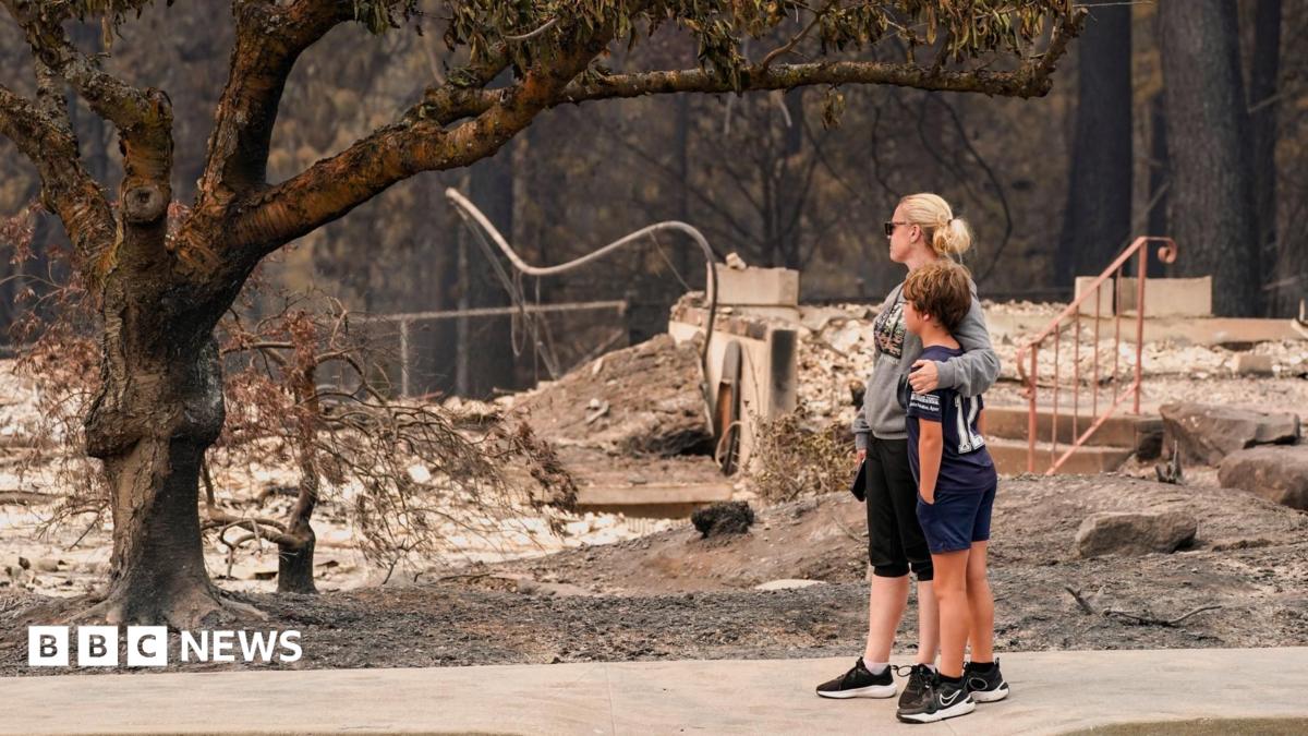 A woman dressed in black capris, grey jumper and black trousers wraps her arm around a young boy in black shorts, black top and black trainers, as both survey the damage of a burnt tree and rubble of a building