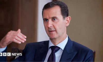 Bashar al-Assad pictured in 2016 speaking while gesturing with one hand. He is wearing a dark suit, white shirt and tie.