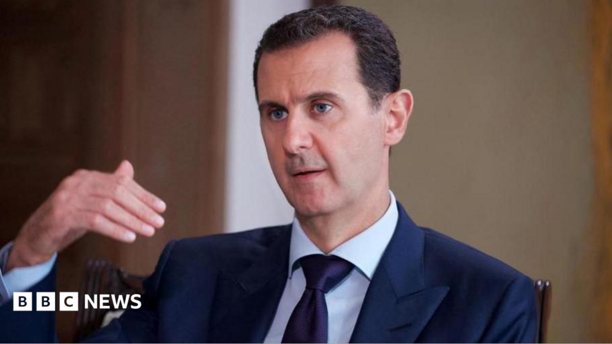 Bashar al-Assad pictured in 2016 speaking while gesturing with one hand. He is wearing a dark suit, white shirt and tie.