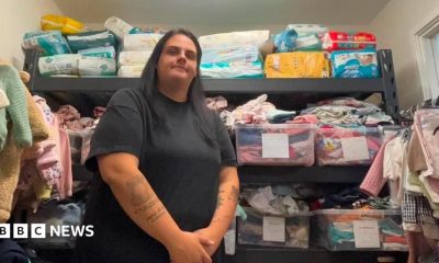 Megan Sinden is standing in front of the baby bank's wares with her hands clasped in front of her and a slight smile on her face. She is wearing a black top and trousers and has long black hair. The wall behind her is stacked with boxes of colourful clothes and nappies. Both walls beside her are also lined with baby clothes hanging on a rails.