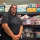 Megan Sinden is standing in front of the baby bank's wares with her hands clasped in front of her and a slight smile on her face. She is wearing a black top and trousers and has long black hair. The wall behind her is stacked with boxes of colourful clothes and nappies. Both walls beside her are also lined with baby clothes hanging on a rails.