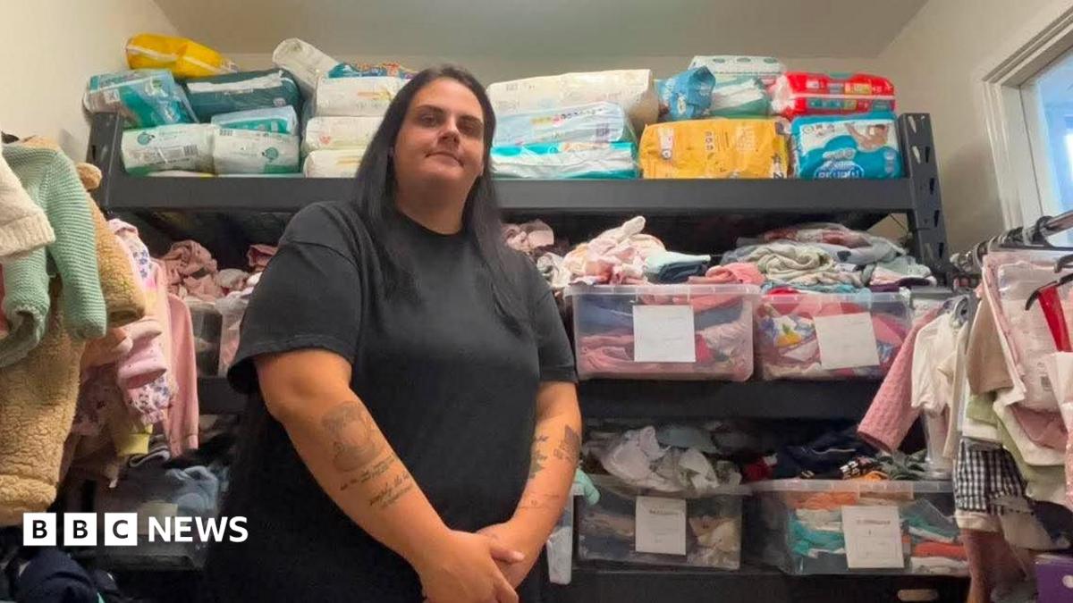 Megan Sinden is standing in front of the baby bank's wares with her hands clasped in front of her and a slight smile on her face. She is wearing a black top and trousers and has long black hair. The wall behind her is stacked with boxes of colourful clothes and nappies. Both walls beside her are also lined with baby clothes hanging on a rails.