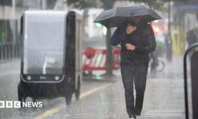A man wearing a sodden jacket and trousers carries an umbrella down a street through a downpour