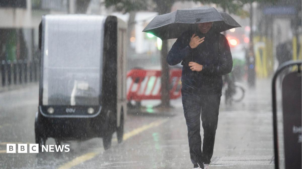 A man wearing a sodden jacket and trousers carries an umbrella down a street through a downpour