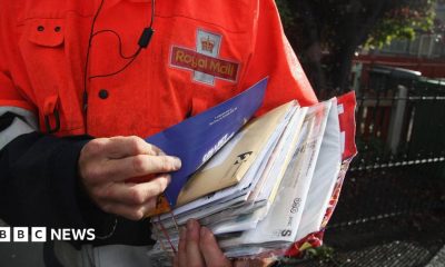 An employee of Royal Mail sorts parcels and letters in the back of his delivery van. The Royal Mail branding is on the back of his jacket and on the side of the van.