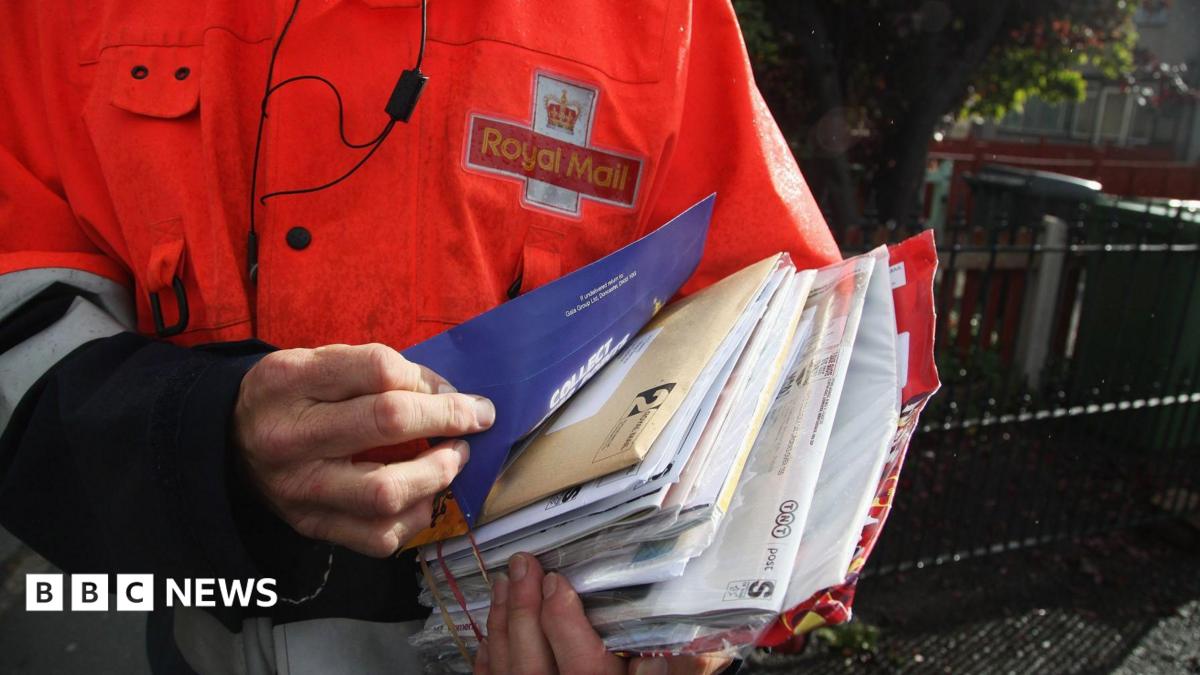 An employee of Royal Mail sorts parcels and letters in the back of his delivery van. The Royal Mail branding is on the back of his jacket and on the side of the van.