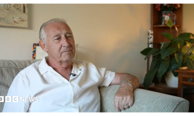 A person with short grey hair sits on a light-coloured sofa in a living room, wearing a white short-sleeved shirt and a wristwatch. In the background, a wooden shelf holds a potted plant and flowers.